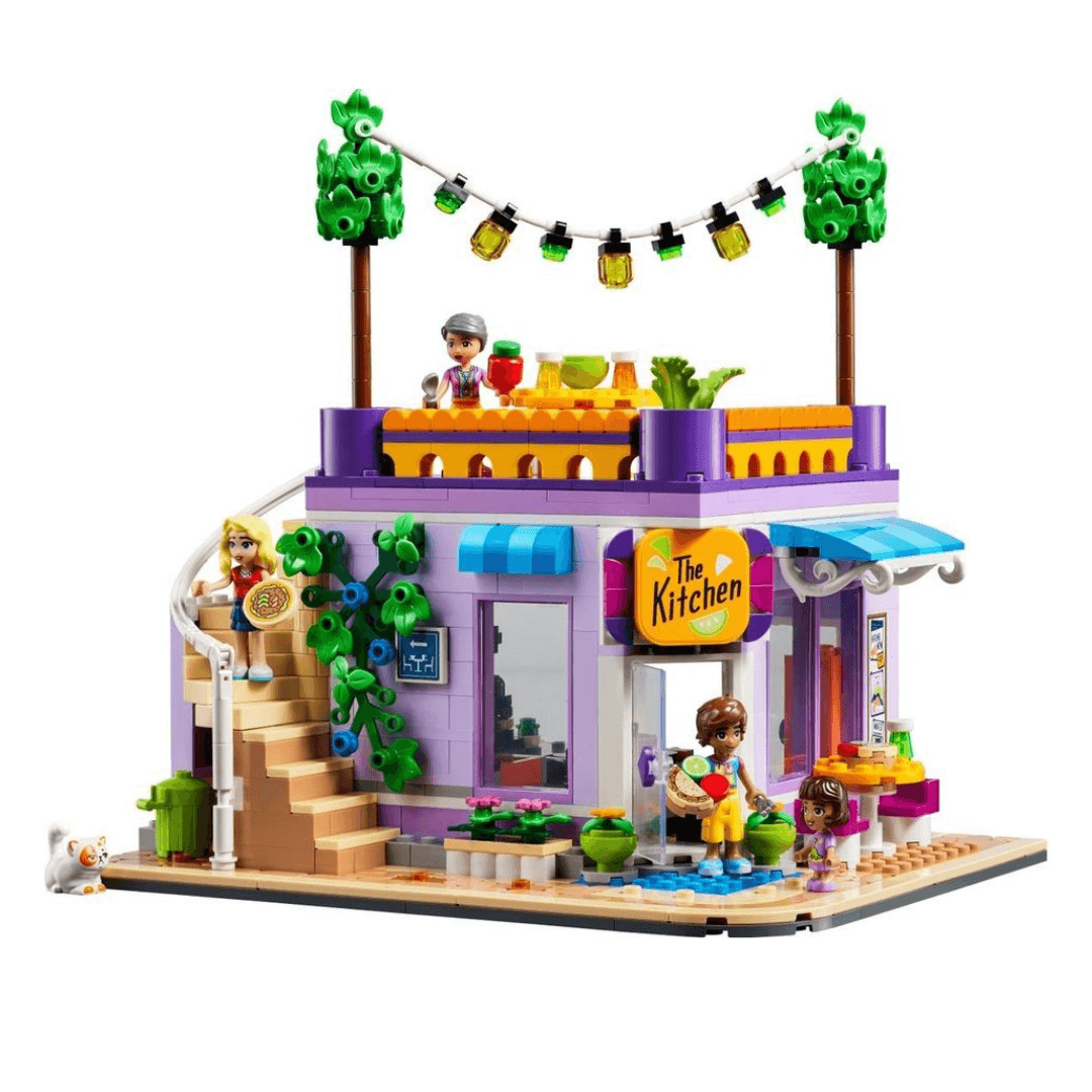 41747 Lego Friends Heartlake City Community Kitchen Built Set. Small Purple room With Large Windows, Stairs Leading Up To A Balcony WIth Small Table And Hanging Lights. Building Surrounded By Small Tables, Vines And Flowers. 4 Characters Included With Fruit, Pizza, Drinks. Small White Cat Sitting In Corner.